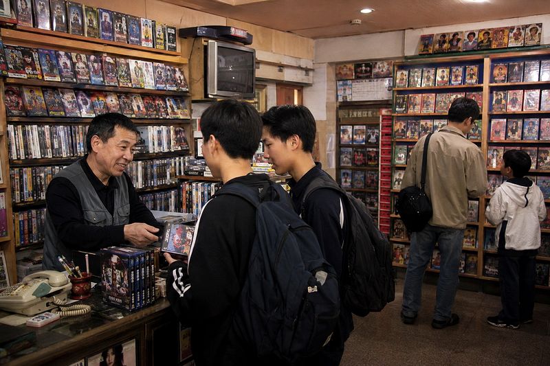 비디오테이프가 가득한 대여점에서 여러 손님이 선반을 살펴보고 주인이 계산대에서 테이프를 건네는 모습