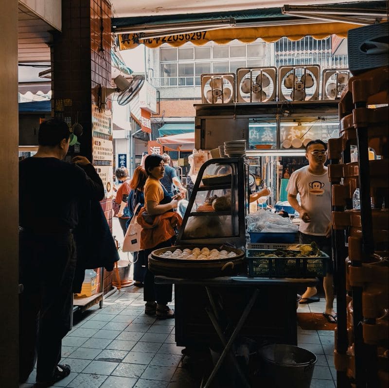 난징의 전통 재래시장 안 작은 식당 입구에서 다양한 재료와 음식이 진열된 모습, 사람들이 북적이며 음식을 주문하고 있는 풍경