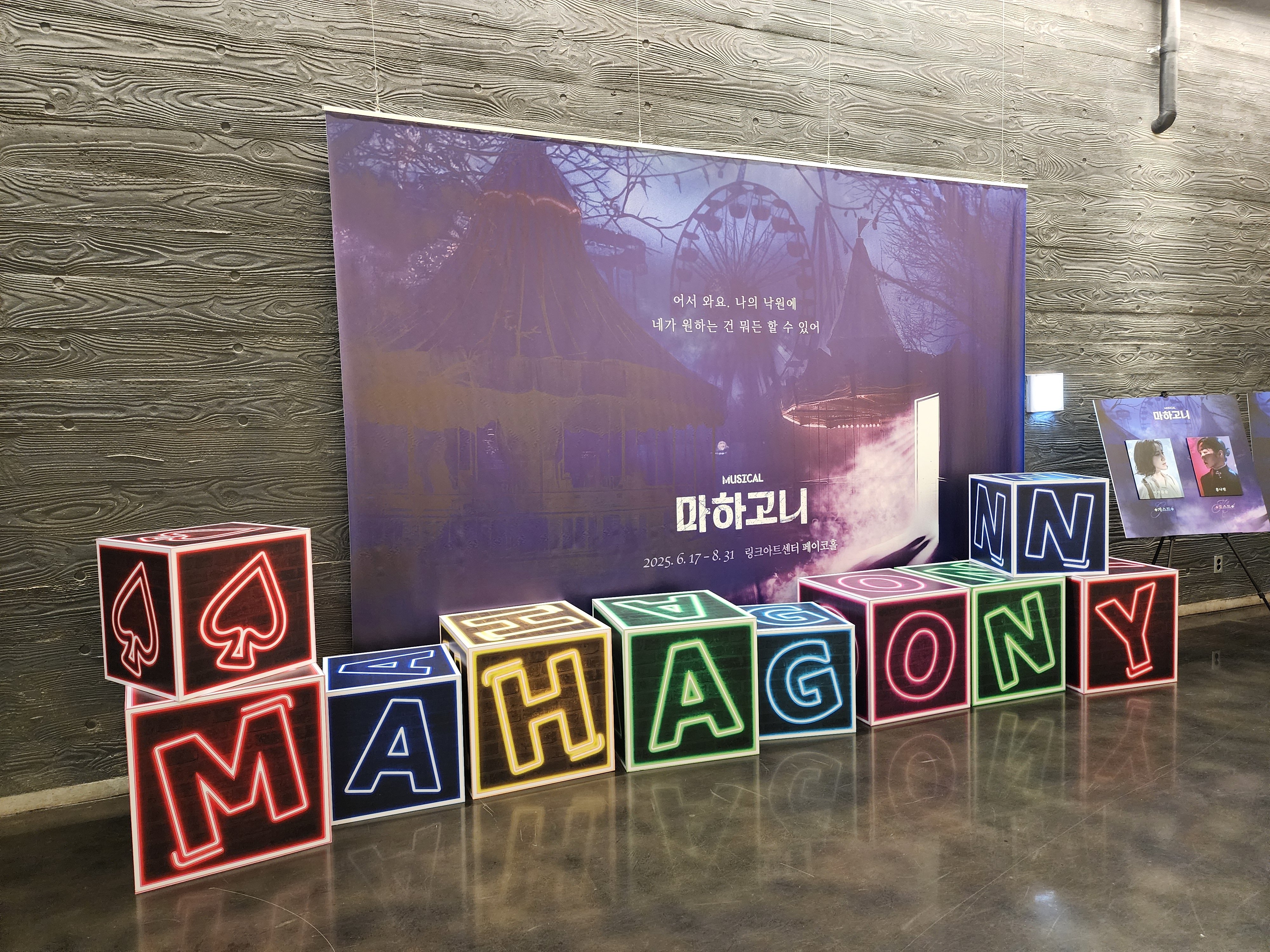 뮤지컬 마하고니 포토존.
폐허가 된 스산한 놀이공원 이미지의 마하고니 메인 포스터를 기반으로 한 현수막 앞에
MAHAGONNY
이니셜이 사각 상자 모양으로 한 글자씩 놓여있음