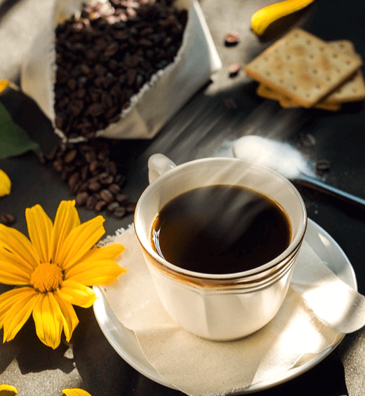 하얀 잔에 검은 블랙커피가 있고 주변엔 노란 꽃 한송이와 커피 원두가 담긴 하얀 자루, 크래커 3개가 있다.