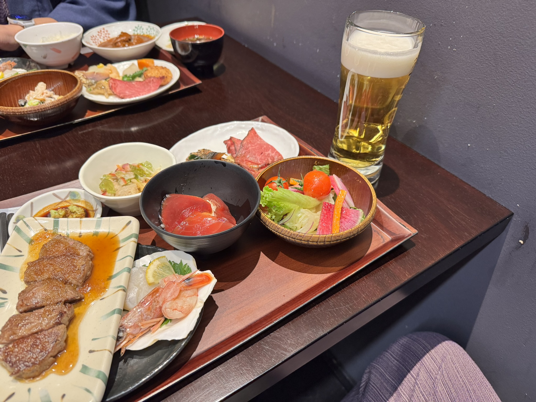 테이블 위에 차려진 풍성한 석식 한 상. 노릇하게 구워진 스테이크, 신선한 참치 회(마구로), 새우 가리비 회, 샐러드, 그리고 시원한 기린 생맥주 한 잔이 놓여 있음.