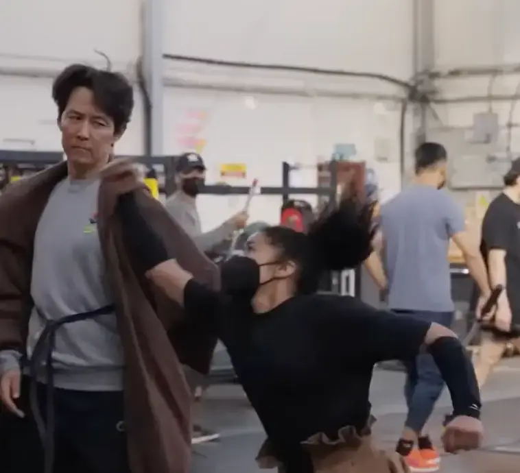 갈색 망토를 두르고 마스크에 검은 티를 입고 달려드는 여자와 무술연습을 하는 이정재