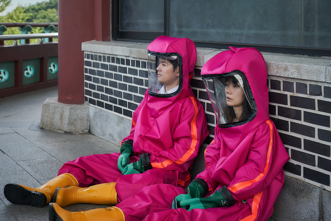 배두나와 장기하가 방호복을 입고 앉아 있다.