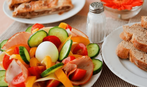 하얀 접시 위에 오이 슬라이스, 햄 슬라이스, 방울토마토들이 가득 하고 삶은 달걀 한 개가 중앙에 있다. 옆 접시엔 호밀식빵이 있다.