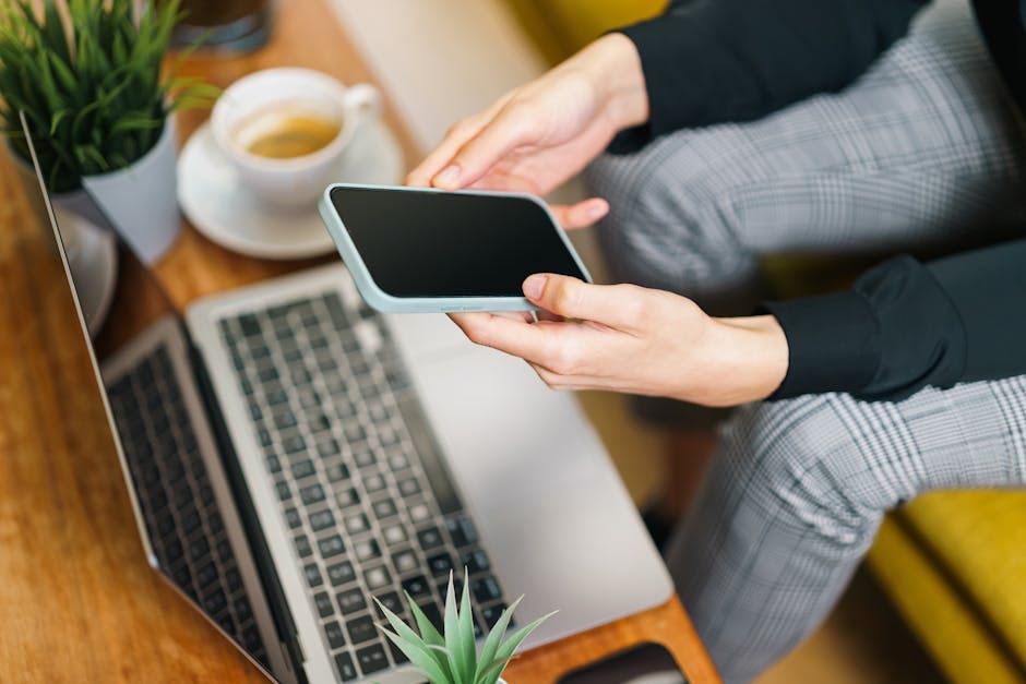 Close-up of hands holding smartphone over a laptop, with coffee and plants on a wooden desk.