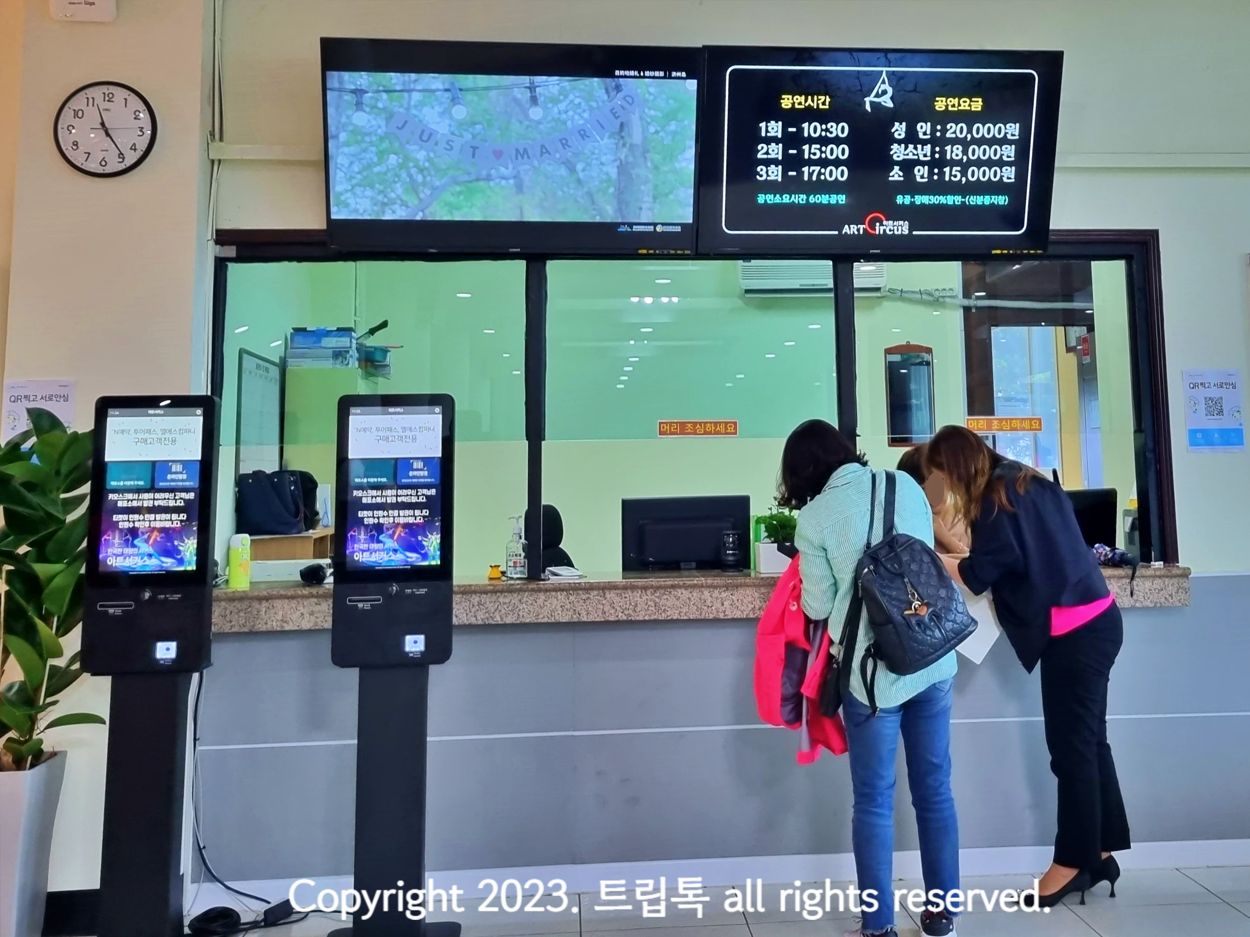 매표소에 2명이 티켓을 구입하고 왼쪽에 키오스크 2대가 있다