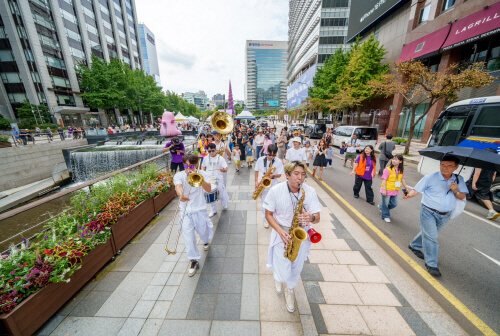 서울 도심 거리에서 브라스 밴드가 퍼레이드 공연을 하며 행진 중인 장면, 시민들과 함께 걷는 거리 축제 분위기