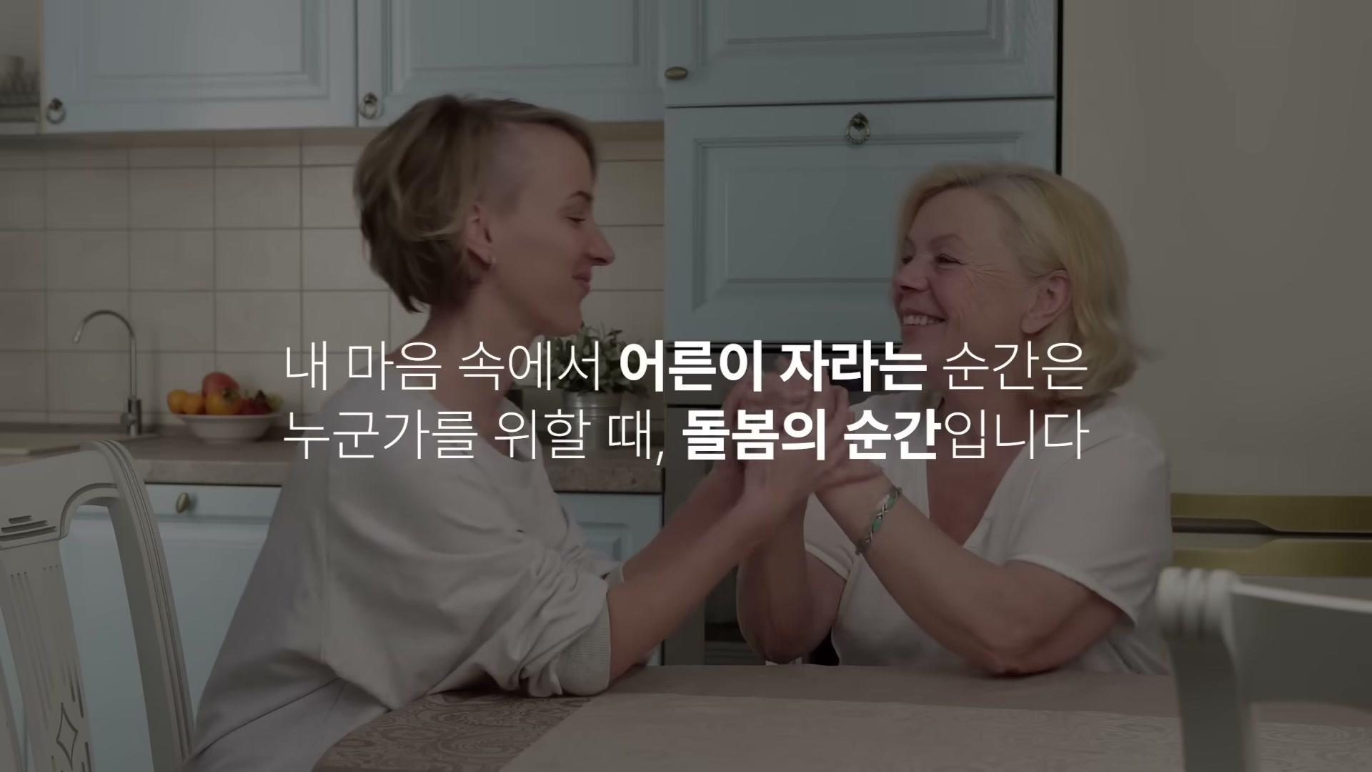 밝은 주방에서 두 여성이 서로 손을 맞잡고 마주 보며 웃는 장면. 화면에 어른이 자라는 순간, 돌봄의 순간 자막이 떠 있다.