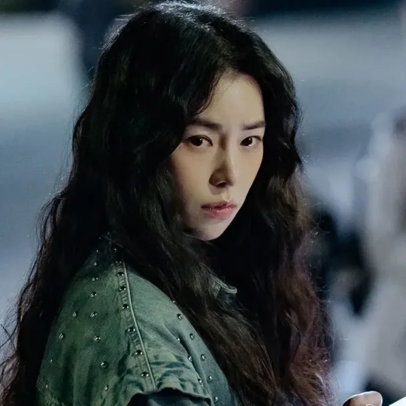 어느 실외를 배경으로 녹색 가죽 점퍼를 입고 긴 파마 머리를 하고 있는 국민사형투표의 주현