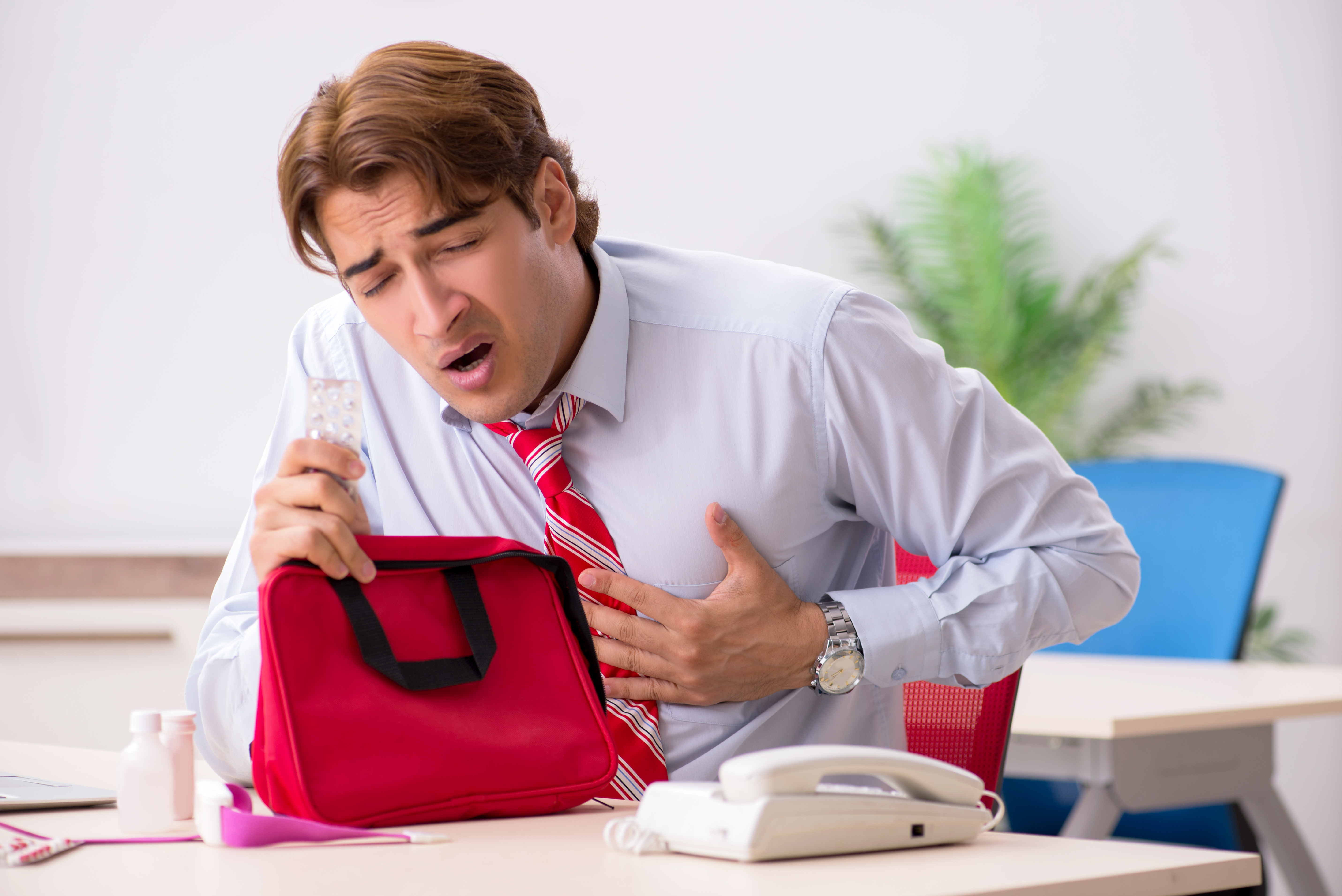 한 남성이 사무실 책상에 앉아 있습니다. 그는 흰색 셔츠와 빨간색 줄무늬 넥타이를 매고 있으며, 왼손으로 가슴을 부여잡고 있습니다. 얼굴은 고통스러워 보이며, 입을 반쯤 벌린 채 숨이 차거나 통증을 느끼는 듯한 표정을 짓고 있습니다. 오른손에는 알약이 든 약 포장을 들고 있으며, 책상 위에는 빨간색 응급처치 가방이 열려 있습니다.