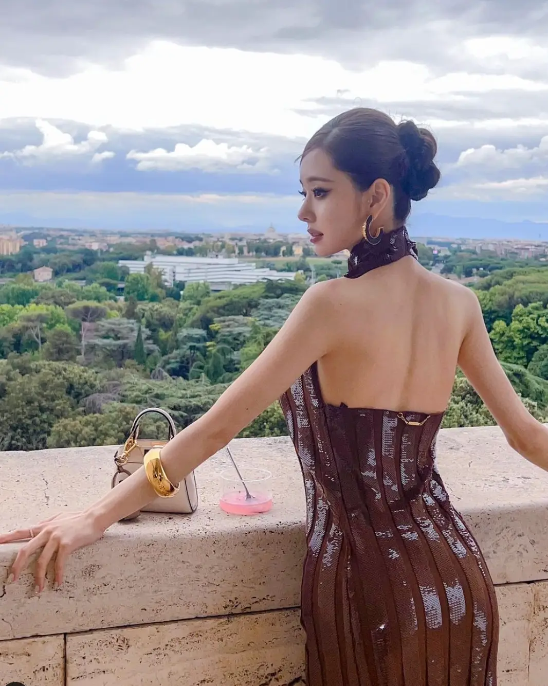 허리춤에 오는 아이보리 돌담을 두손으로 짚고 등이 파인 갈색 몸에붙는 반짝이는 원피스를 입고 머리를 하나로 묶은채 옆을 바라보는 여자의 뒷모습