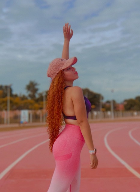 분홍색 그러데이션 레깅스와 보라색 브라톱을 입은 여성이 야외 트랙 위에서 한쪽 팔을 하늘로 뻗으며 포즈를 취하고 있다. 여성은 긴 곱슬 머리에 핑크색 모자를 쓰고 있으며, 배경에는 푸른 하늘과 나무가 보인다.