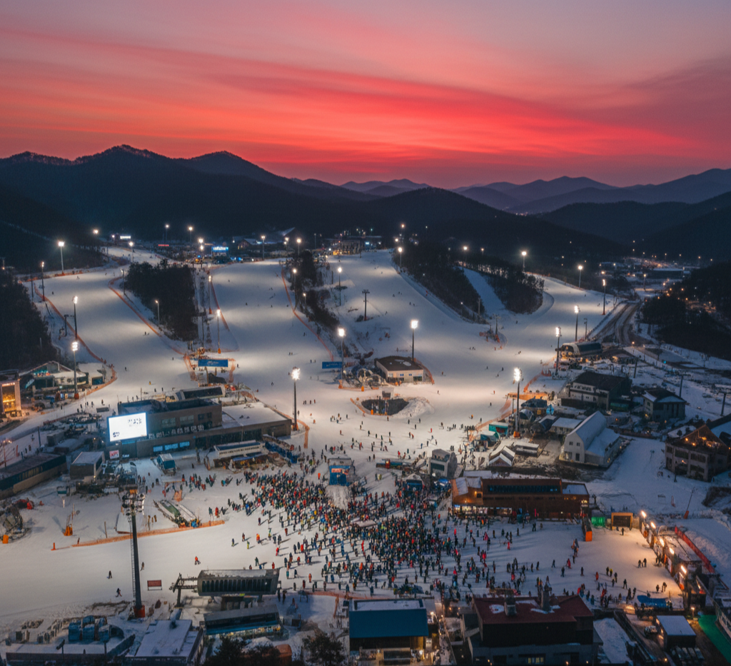 해 질 녘 또는 야간의 강원도 비발디파크 스키장 전경. 환하게 불 밝힌 슬로프 위로 수많은 스키어와 스노보더들이 활동하고 있으며, 멀리 산이 보인다.