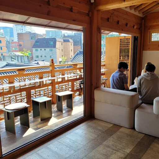 한옥 스타일의 인테리어가 인상적인 서울의 Cafe Sa Sa 내부 모습. 목재 난간 너머로 도심 뷰가 보이고&amp;#44; 창가에는 좌식 좌석과 테이블이 배치되어 있음.