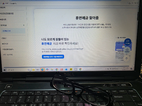 노트북 화면에 띄워진 서민금융진흥원 휴면예금 찾아줌 홈페이지와 그 위에 놓인 60대 주부의 돋보기안경