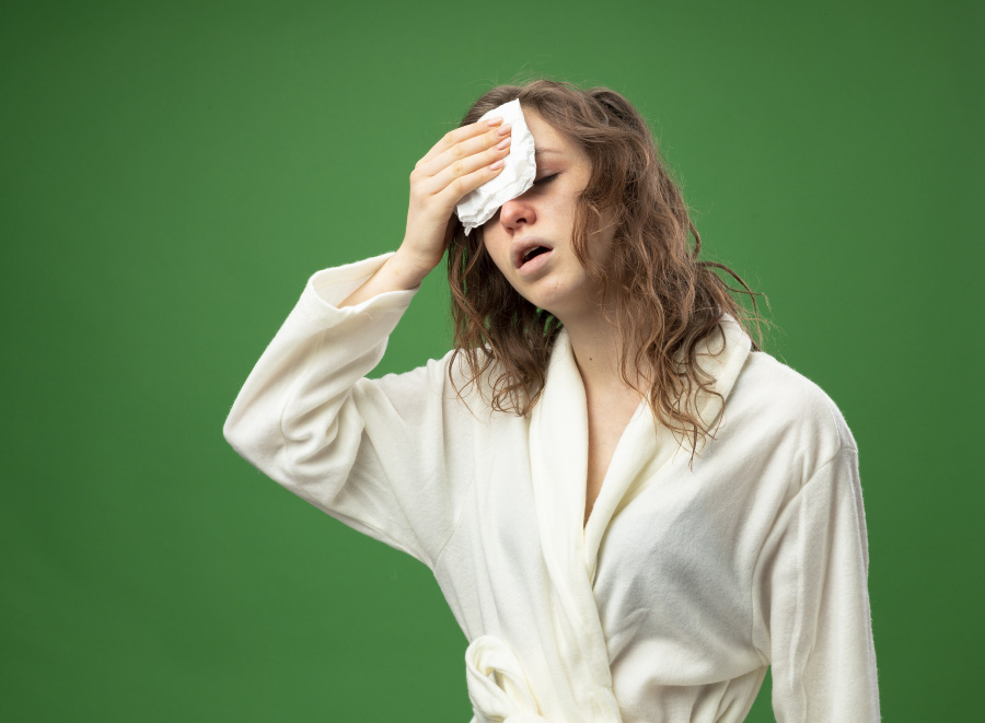 한 서양 여성이 초록색 벽 앞에서 잠옷을 입고 이마에 흐르는 식은땀을 손수건으로 닦고 있는 것을 찍은 사진