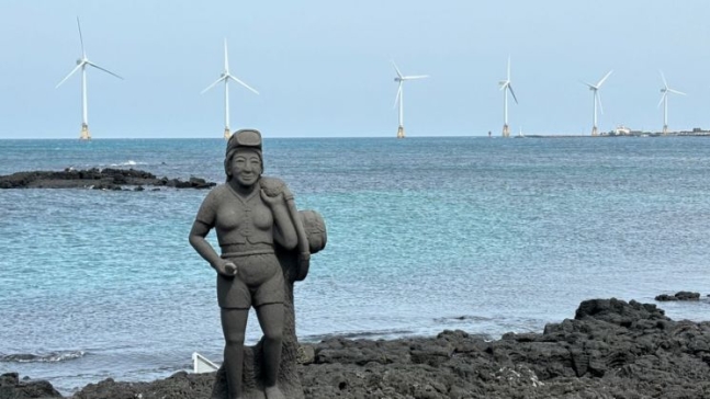 제주도 한경면 해안의 해녀 석상 뒤로 탐라해상풍력발전단지의 풍력 발전기들이 돌아가고 있다.