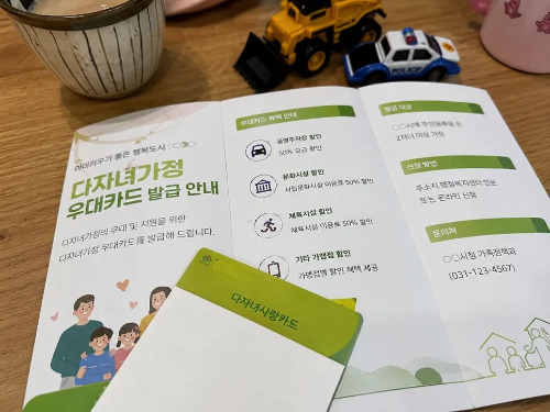 식탁 위에 놓인 다자녀가정 우대카드 발급 안내 리플릿과 다자녀사랑카드, 그리고 뒤쪽에 보이는 장난감 자동차와 커피잔