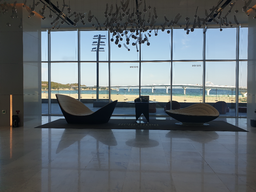 창 밖으로 해변과 바다 그리고 다리가 보인다. 안쪽에는 소파가 놓여 있다.