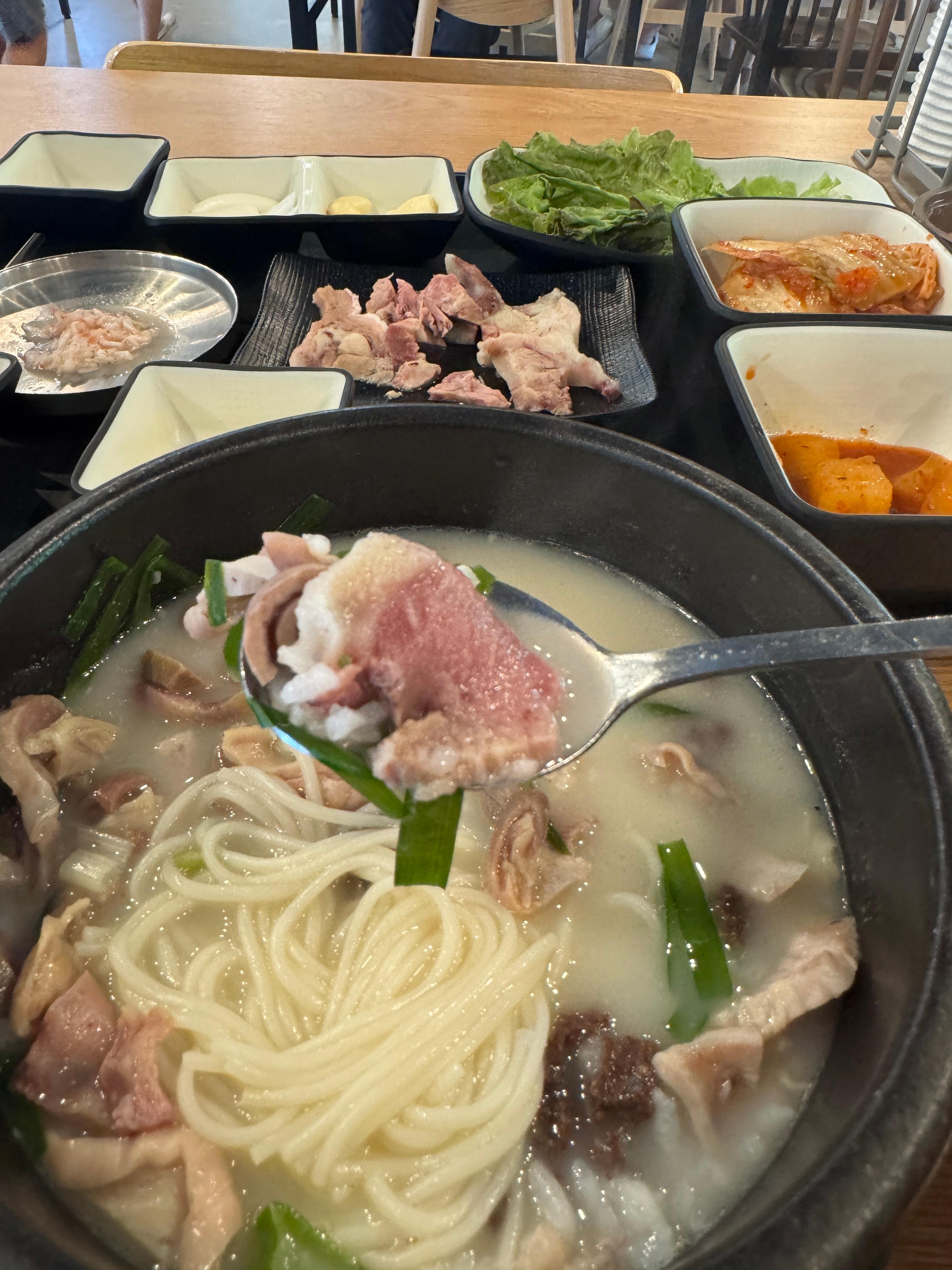 하얀 소면과 고기&amp;#44; 밥이 함께 담긴 숟가락 클로즈업. 국밥 뚝배기 속에서 건져 올렸으며&amp;#44; 뒤로는 수육과 밑반찬이 보인다.