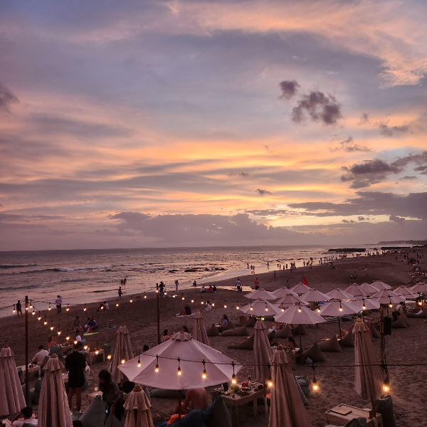 보랏빛 하늘아래 조명과 파라솔 그리고 테이블이 있는 해변이 보인다.