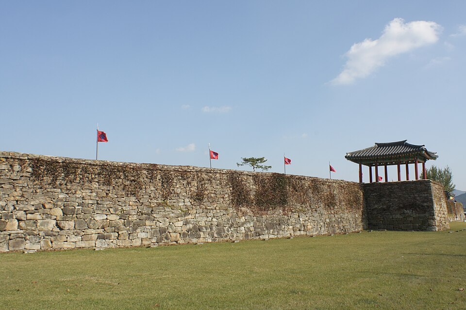 A long stone wall at Haemieupseong leads to an open wooden pavilion, with flags above the wall and grass in front.