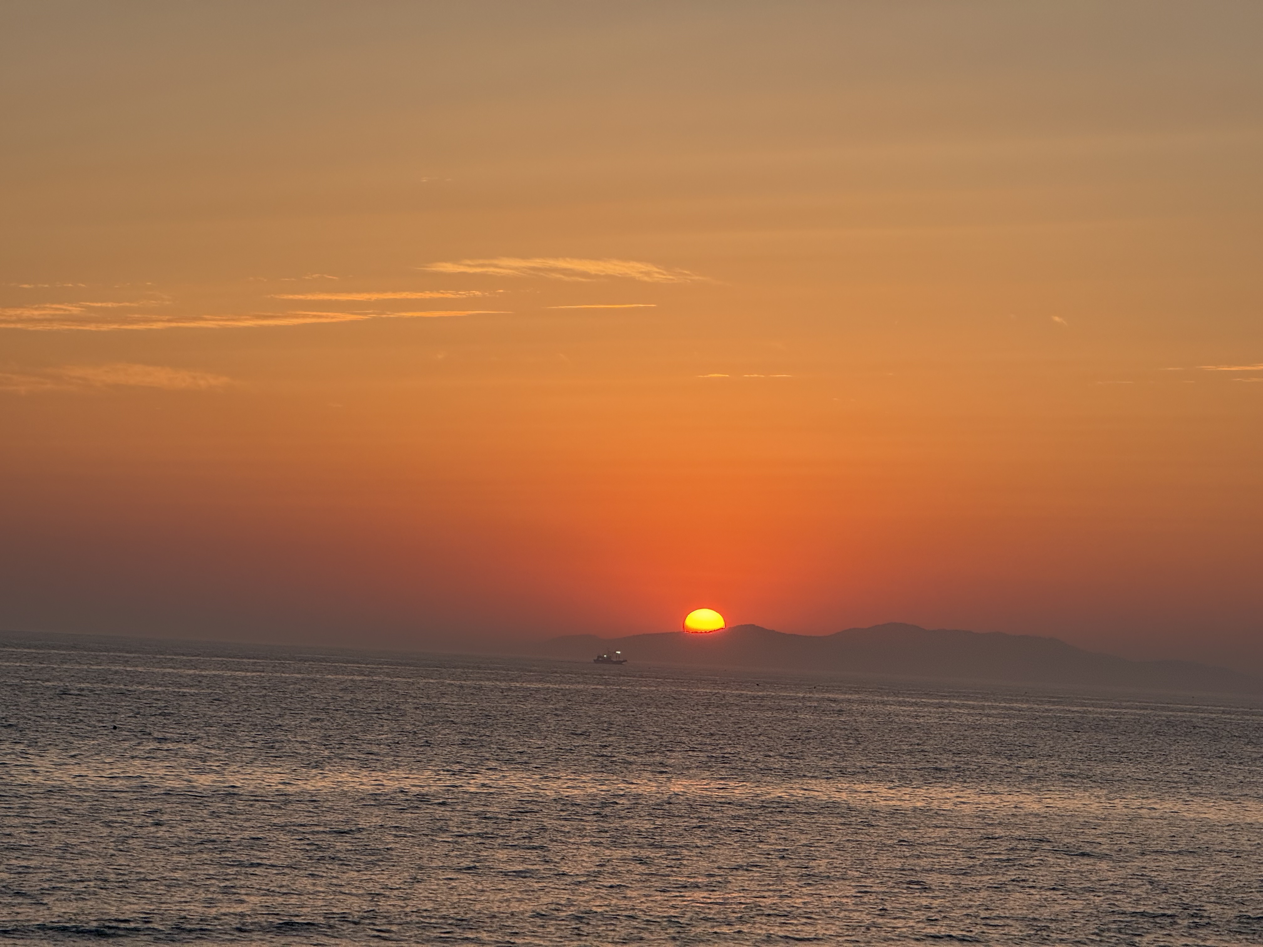 저멀리 배가 있고 바다 너머 보이는 섬 뒤로 해가 절반가량 숨어있는 모습