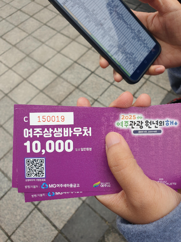 왼손에는 여주 상생 바우처를 오른손으로는 바우처 사용처가 보이는 핸드폰을 들고 있다.