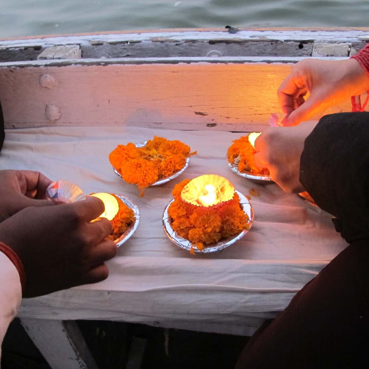 보트 위에서 주황색 꽃으로 장식된 연꽃 등불(디야)에 촛불을 붙이고 있는 사람들의 손