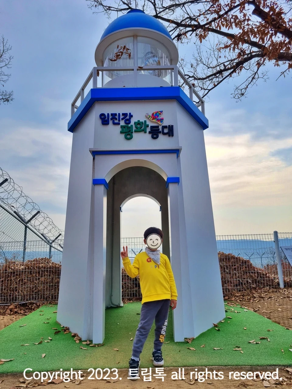 하얀색 길쭉한 건물에 둥그런 파란 기붕을 가진 인진각평화등대 앞에 오란옷을 입은 아이가 서있다.