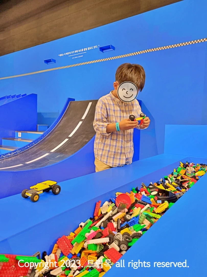 파란색 공간에 검은 경사로가 있고 그 앞에서 아이라 브릭으로 자동차를 만들고 있다