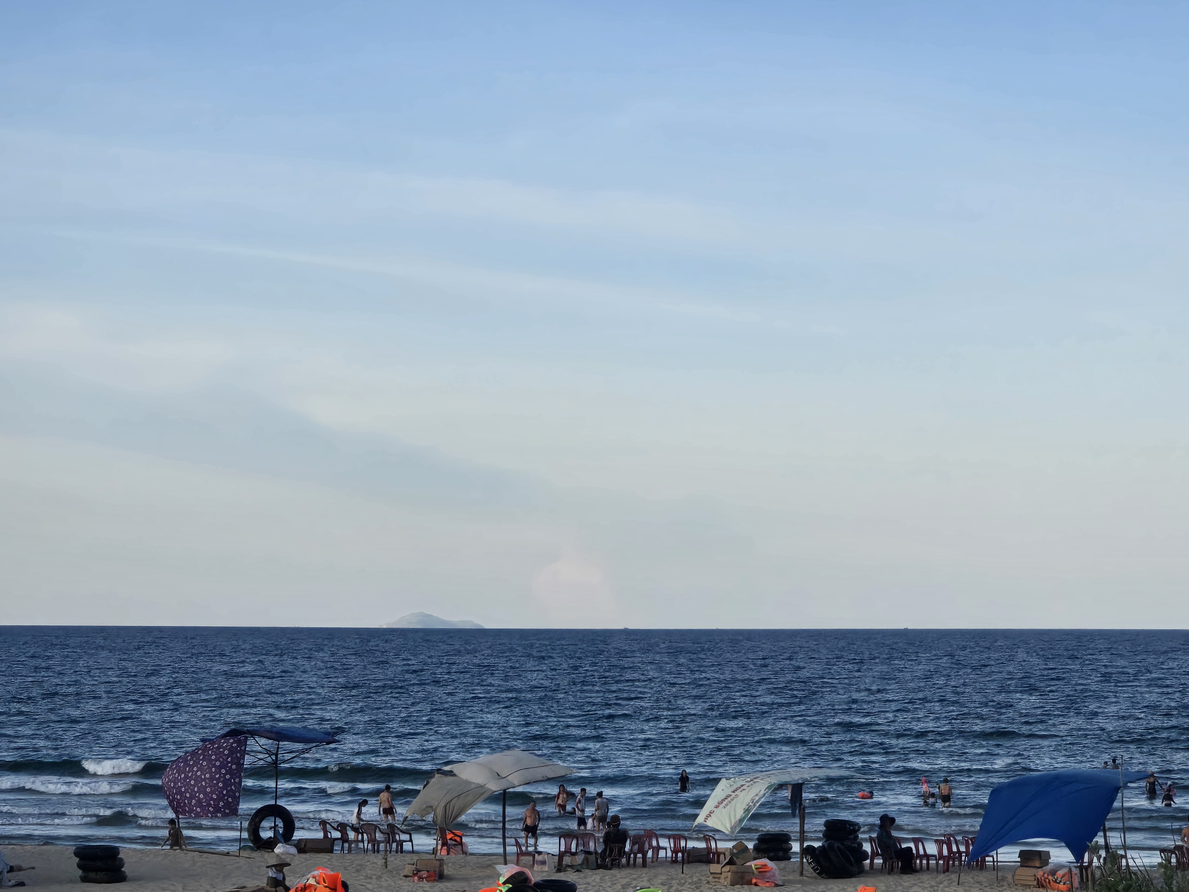 파라솔과 비치체어가 놓인 여름 해변에서 사람들이 잔잔한 파도 속에 물놀이하는 모습, 수평선 너머 작은 섬이 보임