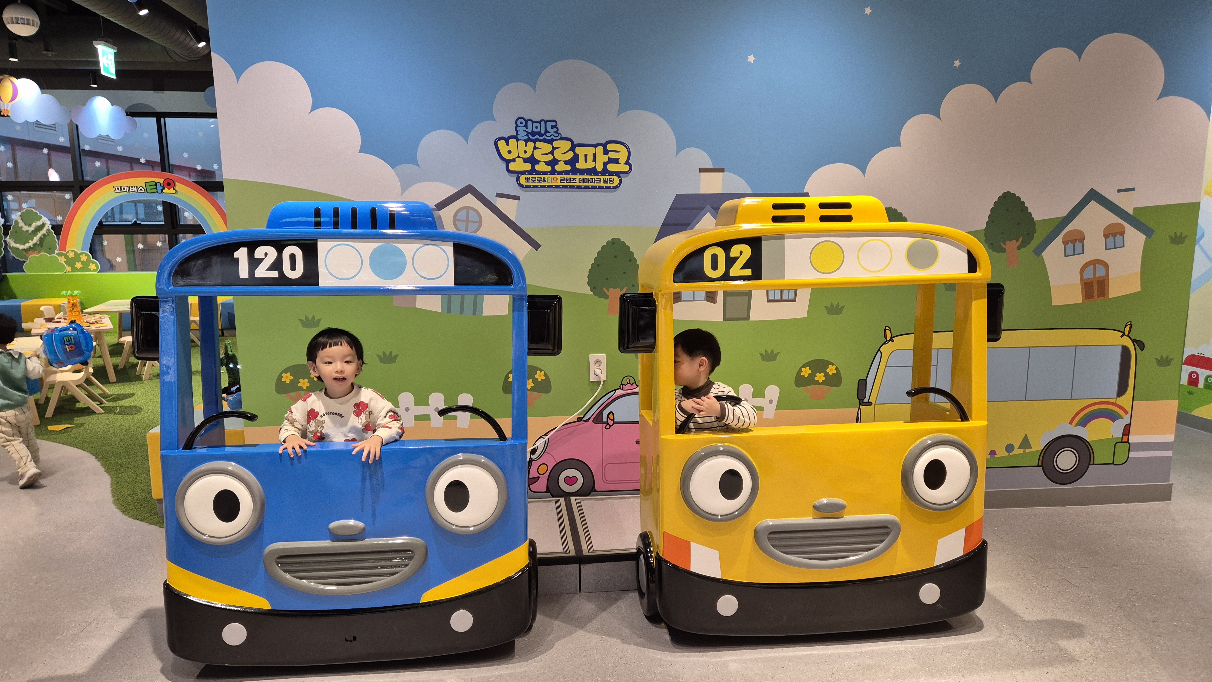 월미도 뽀로로파크에서 파란 타요 버스와 노란 친구 버스를 각각 타고 있는 두 아이의 모습. 아이들의 웃는 얼굴과 알록달록한 배경이 따뜻하고 즐거운 분위기를 전한다.