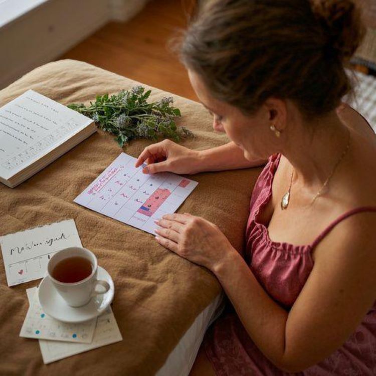 따뜻한 톤의 침실에서 달력을 보며 생리주기를 확인하고 있는 여성의 모습. 한쪽에는 생리 관련 건강 정보가 담긴 노트와 허브차가 놓여있어 자연스럽고 건강한 여성의 일상을 표현하는 이미지