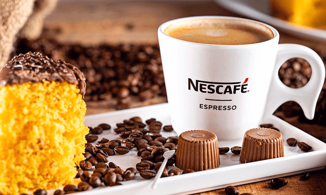 하얀 잔에 카페라떼가 가득하고 왼쪽에는 노란색 작은 빵과 주변엔 커피콩들이 많다.