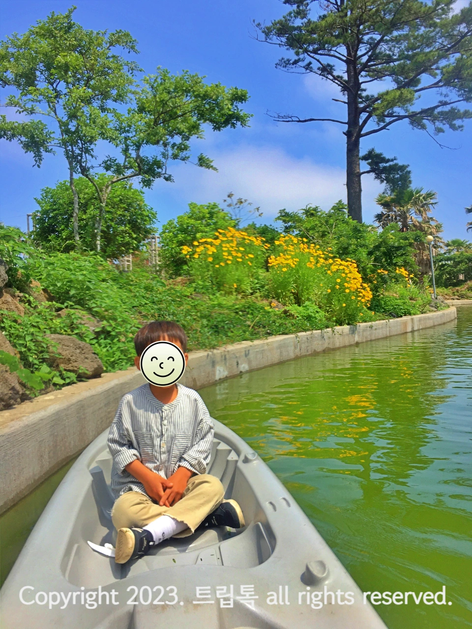 카약에 아빠다리 하고 앉은 아이 뒤로 초록색 물과 노란 꽃이 보인다