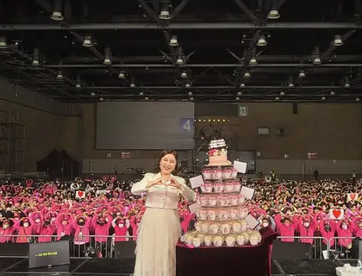 송가인- 실내 콘서트장 가운데 흰옥을 입고 두손으로 하트를 만들며 웃는 송가인 여 팬들이 만들어준 엽서를 케이크 모걍으로 쌓은 탑 뒤 분홍색 옷을 입고 두손으로 머리하트를 만즐고 있는 팬들