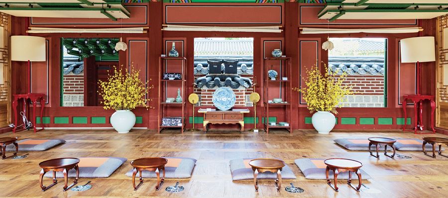 경복궁 생과방 체험장. 나무로 된 바닥에 1인 둥근 상이 7개와 함께 방석이 준비되어 있으며, 인테리어로는 노란색의 꽃이 꽂힌 화병 2개가 있고 그 뒤로 난 창문 3개를 통해 건물 외부의 기와로 덮힌 담벼락이 보인다.