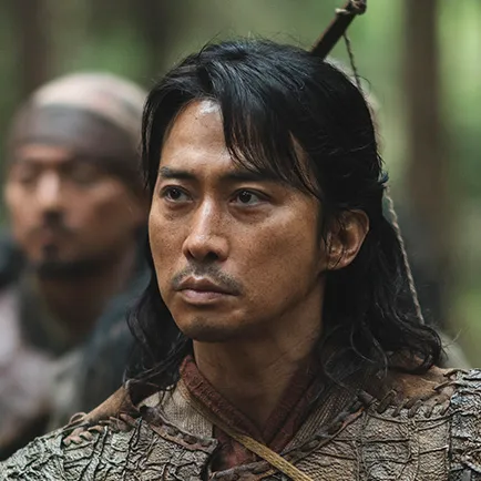 병사를 배경으로 30대 남성이 긴 머리를 뒤로 묶고 허름한 갈색 가죽을 갑옷처럼 두르고 있는 아라문의 검 태마자 캐릭터