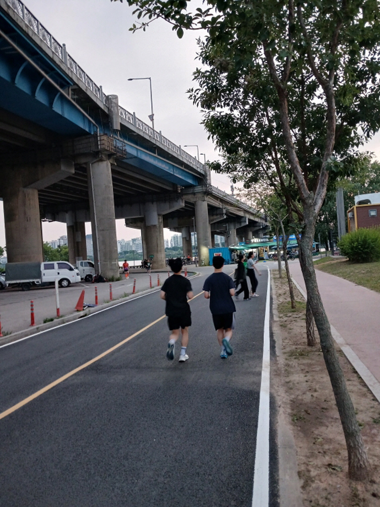 two young boys jogging hard on asphalt under a highway bridge – symbolizing overexertion at the start of exercise./고가도로 아래 아스팔트에서 무리하게 조깅하는 어린 소년들 – 운동 초기에 무리한 운동을 상징함.