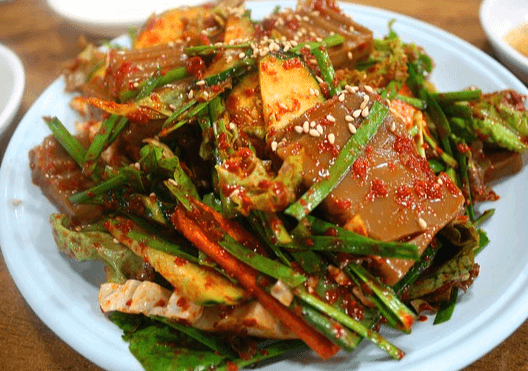 하얀 접시에 도토리묵무침이 담겨 있다. 사각형의 도토리묵이 슬라이스로 잘라졌고 부추와 고추가루 양념장으로 버무린 무침이다.