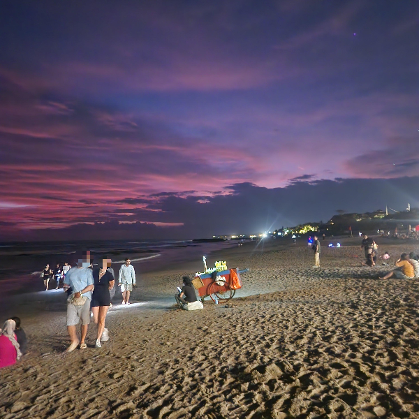 보라색으로 변한 하늘. 어둠이 내리기 시작한 해변이지만 조명으로 인해 밝다. 사람들이 걷거나 앉아 있다.