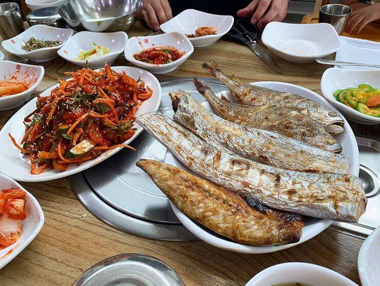 사진 오른쪽에는 빨간 양념과 야채&#44; 서대회가 함께 무쳐진 서대회무침이 접시에 담겨있고 왼쪽에는 접시 위에 구은 고등어&#44; 갈치2토막&#44; 조기 2마리가 있다. 그리고 서대회와 생선구이 주변에 밑반찬들이 있다.