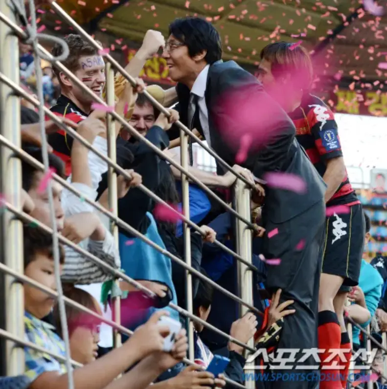 황선홍- 꽃가루가 날리는 축구경기장에서 양복입은 황선홍 감독이 축구관중을 안아주며 웃는모습