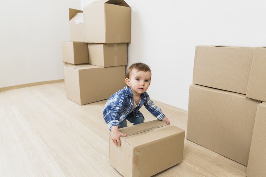 방에 상자가 여러 개 쌓여 있고, 그 중에 하나를 서양 어린 아이가 양쪽 손으로 번쩍 들려고 하고 있는 것을 찍은 사진