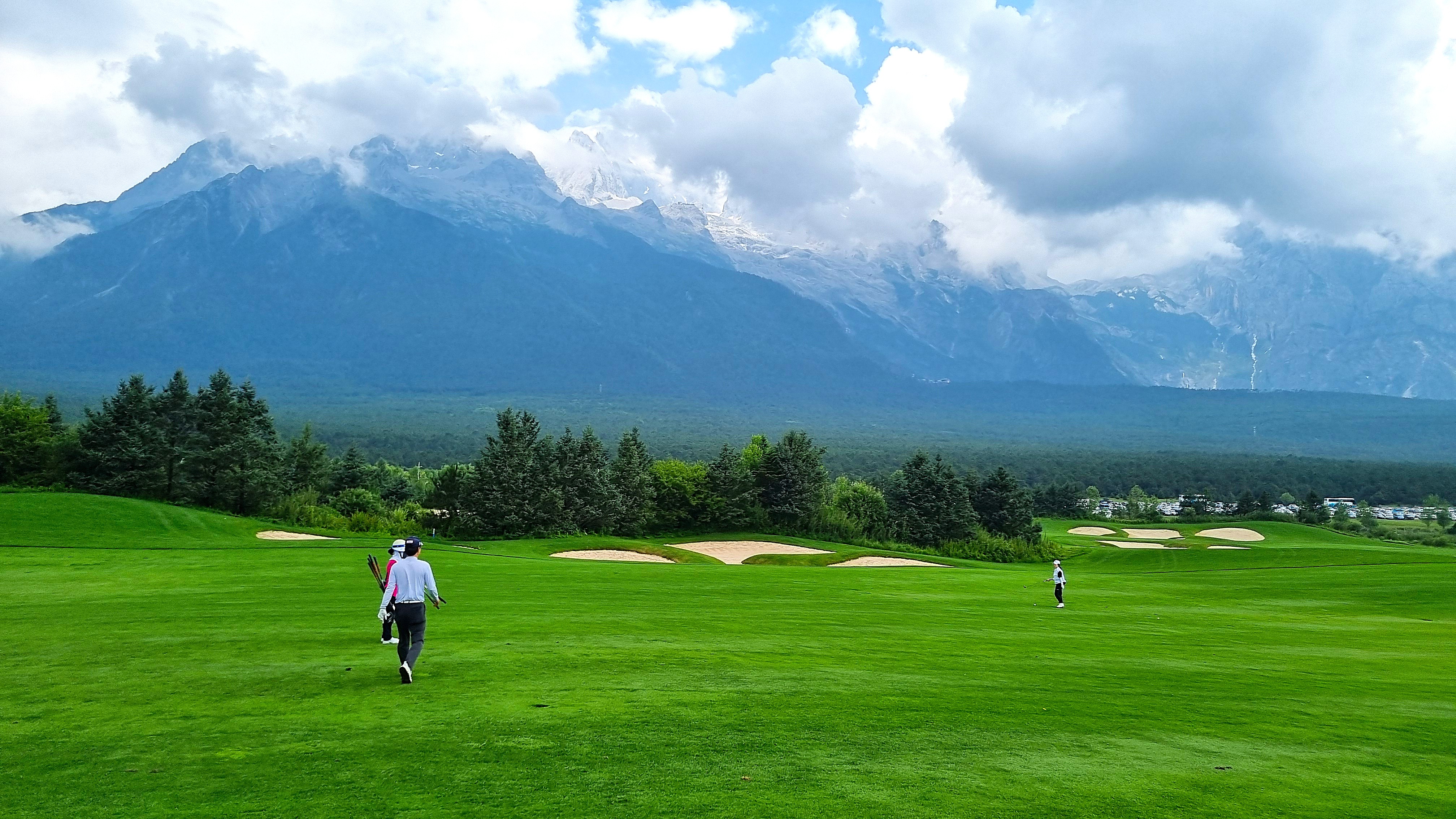 중국 여강 옥룡설산 골프장의 광활한 페어웨이를 따라 걷는 골퍼와 캐디. 배경에는 장엄한 옥룡설산과 구름이 어우러진 절경이 펼쳐진다.