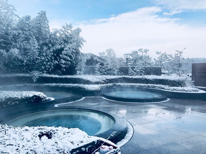 파라다이스 스파 도고의 야외 온천이 둥근 모양으로 가족규모 크기의 온천장이 띄엄띄엄 있고 주변의 나무는 눈으로 덮혀있고 온천장위로는 김이 모락모락 피어오르는 사진