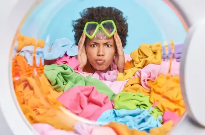 형형색색의 빨래 더미에 얼굴을 묻고 세탁기를 바라보는 고글 쓴 여성의 당황한 표정