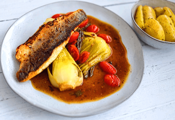 하얀 접시에 농어구이 반마리가 있고 옆에는 볶은 청경채와 방울토마토가 있다. 옆 그릇엔 노란 감자조림 반찬이 있다.