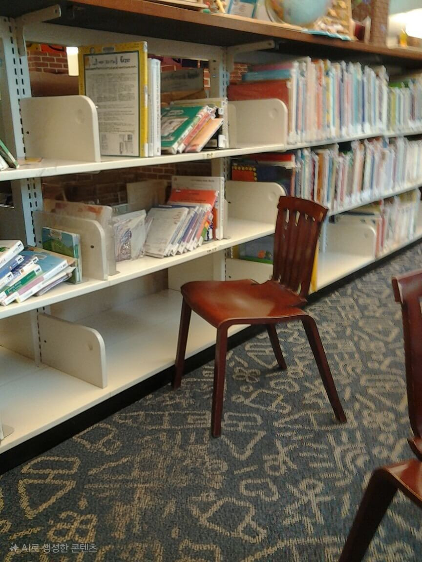 어린이들의 키에 맞춘 낮은 흰색 책꽂이에 알록달록한 그림책들이 가득 꽂혀 있다. 서가 앞에는 아이들이 앉아서 책을 볼 수 있는 작은 나무 의자가 놓여 있으며, 바닥에는 귀여운 그림 패턴의 카펫이 깔려 있다.